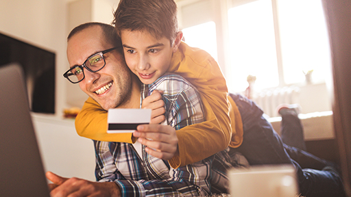 A father sits at a computer wearing glasses. A young child leans over his shoulders from behind holding a credit card.