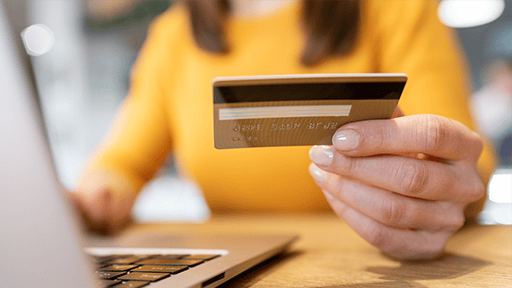A woman in a yellow sweater holds a credit card while using a laptop.