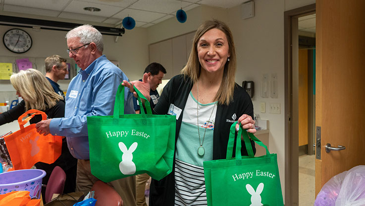 An adult faces the camera smiling holding two green bags with a white bunny and "happy easter" written on the front. 