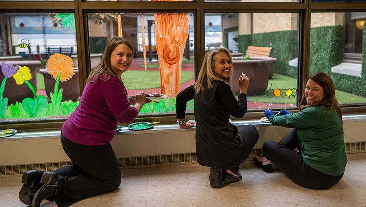 Three adult volunteers paint a window. They are turned toward the camera holding paint brushes. 