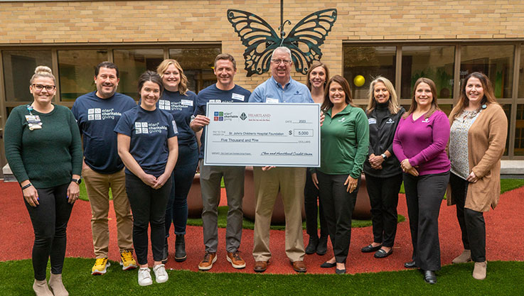 A group of adults stand in an outdoor play area and garden holding a promotional check. 