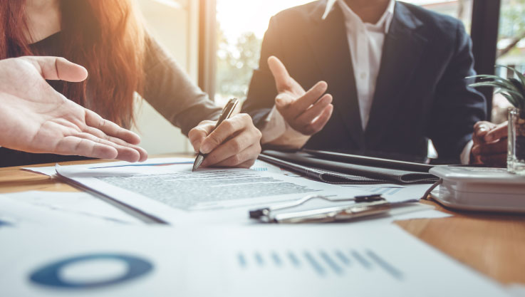 Three people sit around a table covered in papers. The person in the middle is signing a document. 